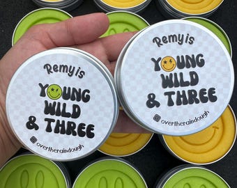 May include: A set of eight small, round containers with lids. Each container holds a bright yellow or green playdough with a smiley face design. The lids are labeled with the text "Remy is Young Wild & Three" and a social media handle.