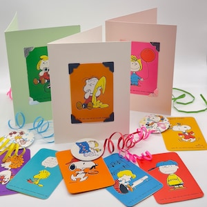 3 cards with vintage peanuts cards attached. very bright, bold colours - orange, blues, red, purple, yellow and pink. Playing cards each feature either Snoopy, Charlie, Lucy, Woodstock, Linus, sally, Freda, schroeder. Card is shiny pink or yellow.