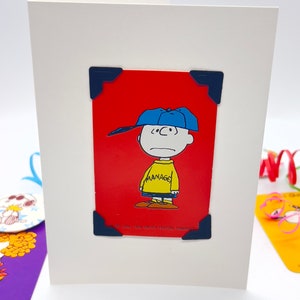 White card with red playing card featuring Charlie Brown with his baseball cap on and his classic yellow jumper with word Manager across it.