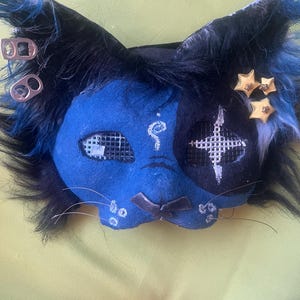 May include: A cat-shaped mask featuring a blue and black face with faux fur ears, mesh eye coverings, and silver soda tab accents. The mask includes three yellow star embellishments, a white spiral symbol on the forehead, and thin whiskers.