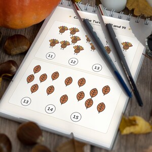 Pode incluir: Uma folha de exercícios educativa encadernada em espiral com atividades de contagem ilustradas com perus e folhas. A página inclui o texto Count the Turkeys 12 and 14, juntamente com círculos numerados 11, 12, 13, 14 e 15. Duas canetas escuras repousam sobre o papel.