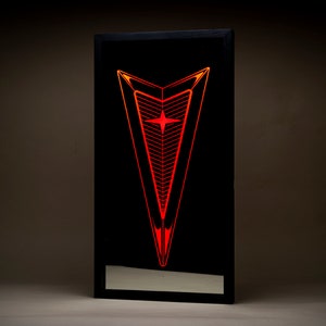 May include: A black framed mirror with a red and orange illuminated Pontiac logo. The logo is a stylized arrow with a star in the center.
