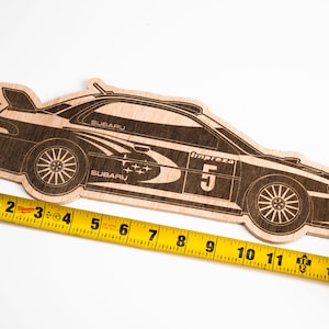 May include: A wooden cutout of a Subaru Impreza rally car, painted black with white and blue accents. The car is numbered "5" and has the word "Impreza" written on the side. The car is being measured with a yellow tape measure, which shows that it is approximately 11 inches long.
