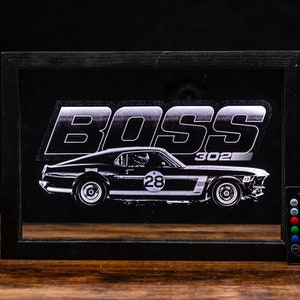 May include: A black and white image of a Ford Mustang Boss 302 with the word "BOSS" above it. The car has a number "28" on the side. The image is in a black frame with a remote control for a light.