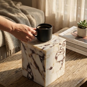 May include: A square-shaped side table made of white marble with prominent dark brown and black veining. The table features a smooth, polished surface and a solid, block-like structure. A matte black ceramic mug filled with coffee sits on top.