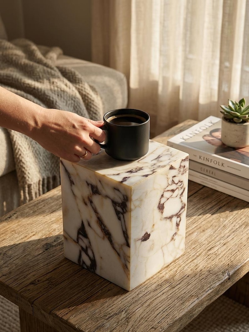 May include: A square-shaped side table made of white marble with prominent dark brown and black veining. The table features a smooth, polished surface and a solid, block-like structure. A matte black ceramic mug filled with coffee sits on top.