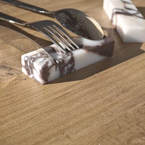May include: Marble utensil rests with a white and brown veined pattern. A fork and spoon rest on one, while another is empty. The rests are rectangular and sit on a wooden surface. The utensils are stainless steel.