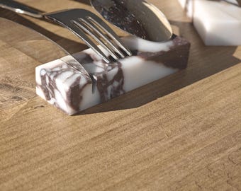 May include: Marble utensil rests with a white and brown veined pattern. A fork and spoon rest on one, while another is empty. The rests are rectangular and sit on a wooden surface. The utensils are stainless steel.