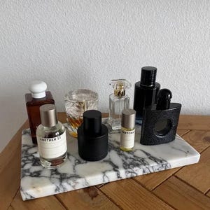Pode incluir: Uma coleção de sete frascos de perfume de várias formas, tamanhos e cores, incluindo vidro transparente, preto e âmbar, dispostos numa bandeja retangular de mármore branco com veios cinzentos. Os rótulos incluem Another 13 e Matcha.