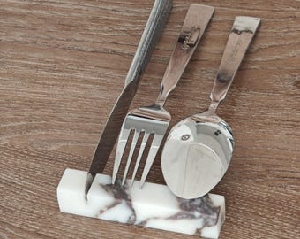 May include: A stainless steel knife, fork, and spoon set resting in a white and gray marble utensil holder. The silverware is arranged on a wooden surface, showcasing a modern dining aesthetic.