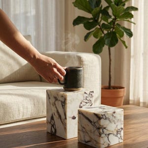 May include: Two square marble blocks featuring white stone with dark purple and black veining. One block is taller than the other, and a dark ceramic mug with visible steam is positioned on top of the taller marble block, showcasing a natural stone finish.