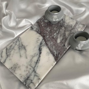 May include: A white marble serving tray with gray veining, featuring two gray ceramic candle holders. The tray is on a white satin fabric background.