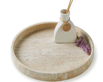 May include: A round, beige travertine tray with a white ceramic diffuser bottle and a sprig of purple flowers. The diffuser bottle has a wooden tag tied around the neck.