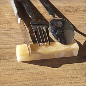 May include: A stainless steel knife, fork, and spoon set resting on a rectangular, cream-colored onyx holder. The utensils are arranged on a wooden surface, with the knife positioned in the holder.