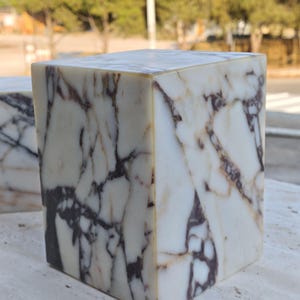May include: A rectangular block made of white marble featuring prominent dark purple and brown veining throughout its surface. The stone object has sharp, clean edges and a smooth, polished finish that highlights the natural patterns of the material.