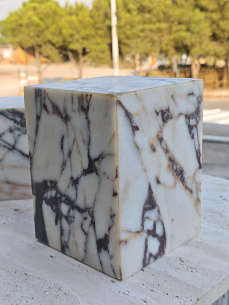 May include: A rectangular block made of white marble featuring prominent dark purple and brown veining throughout its surface. The stone object has sharp, clean edges and a smooth, polished finish that highlights the natural patterns of the material.