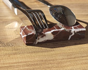 May include: A stainless steel fork and spoon rest on a rectangular marble holder with red and white veining. The utensils are placed on a wooden surface, creating a simple yet elegant table setting.