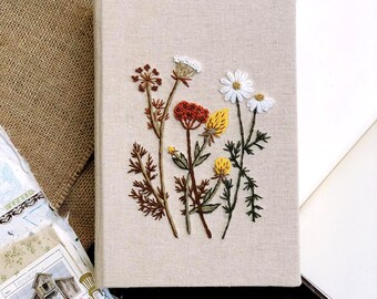 May include: A linen journal cover with embroidered wildflowers in shades of white, yellow, orange, and brown. The flowers are arranged in a cluster in the center of the cover.