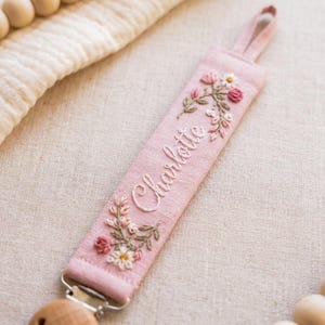 May include: A light pink fabric pacifier clip featuring the embroidered name Charlotte in white cursive script, accented with floral embroidery in shades of pink, white, and green, and finished with a wooden clip and a fabric loop.
