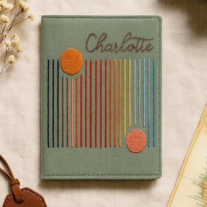 Könnte beinhalten: Salbeigrüner Reisepasshalter mit gesticktem Design. Mit dem Namen "Charlotte" in Schreibschrift, farbigen vertikalen Linien und zwei orangefarbenen Kreisen. Ein Reiseaccessoire.