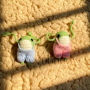 Pode incluir: Dois peluches de sapo de crochê vestindo macacões. Um sapo está vestindo um macacão azul e o outro está vestindo um macacão rosa. Os sapos estão sentados em uma superfície felpuda marrom claro.