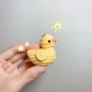 May include: A small yellow crocheted chick with black eyes and an orange beak. The chick is being held in a hand against a white background. A simple drawing of a sun with a smiley face is in the upper right corner of the image.