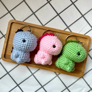 Puede incluir: Tres llaveros de dinosaurio de crochet en azul, rosa y verde. Cada llavero tiene ojos negros y un pequeño anillo de metal.