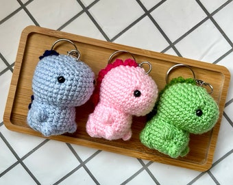 May include: Three crocheted dinosaur keychains in blue, pink, and green. Each keychain has black eyes and a small metal ring.