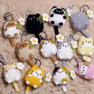May include: A collection of crocheted cat keychains in various colors, including black, white, brown, yellow, and pink. Each keychain features a small crocheted cat with a flower charm attached.