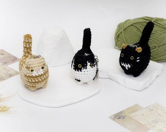 May include: Three crocheted cat keychains. One is black and white, one is brown and white, and one is black. They are all sitting on a white surface.