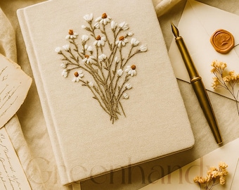 May include: A cream-coloured journal with a floral embroidery, a gold pen, envelopes with wax seals, and a small glass bottle with a cork. The items are arranged on a cream fabric surface.
