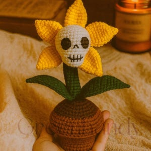 May include: Crocheted flower with yellow petals and a skull face, green leaves, and a brown pot. The handmade craft is held in a hand, with a lit candle in the background. The flower is a unique decorative item.