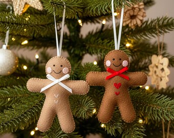 May include: Two felt gingerbread man ornaments hanging on a Christmas tree. One is light brown with a white bow tie, and the other is dark brown with a red bow and heart. The tree is decorated with gold and white ornaments.