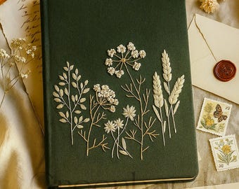 Könnte beinhalten: Ein dunkelgrünes Notizbuch mit gestickten Blumenmustern in Creme und Gold. Das Notizbuch ist von Schreibwaren umgeben, darunter Umschläge mit Wachssiegeln, Briefmarken und einem Vintage-Stift, was eine Vintage-Ästhetik erzeugt.