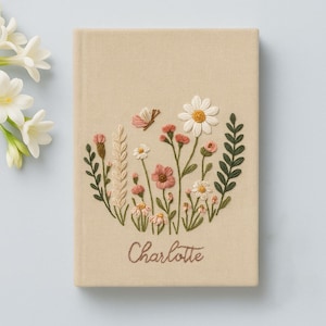May include: A beige linen journal with a floral embroidery design. The embroidery features pink and white flowers, green leaves, and a butterfly. The name "Charlotte" is embroidered in cursive below the flowers.
