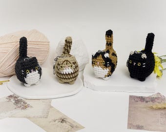 May include: Four crocheted cat keychains in different colors and patterns. The cats are sitting on a white surface.