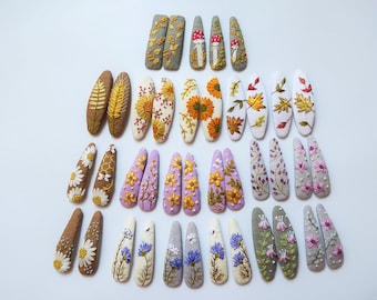 Può includere: Una collezione di fermagli per capelli ricamati con vari disegni floreali e ispirati alla natura. I fermagli presentano ricami dettagliati di girasoli, margherite, foglie e funghi in una varietà di colori, tra cui giallo, arancione, bianco e viola. I fermagli sono disposti su una superficie bianca.