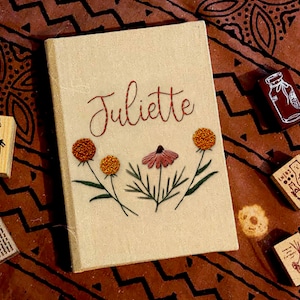 May include: A beige linen journal with the name "Juliette" embroidered in brown thread. The journal is decorated with embroidered flowers in shades of orange and pink.