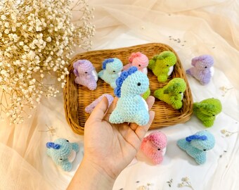 May include: A collection of colorful, crocheted dinosaur toys in a wicker basket. The dinosaurs are in various pastel colors, including blue, green, pink, and purple. One light blue dinosaur with a blue crest is held in a hand. The background is a soft, neutral fabric.