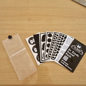 Könnte beinhalten: Ein schwarz-weißes Reise-Set mit LED-Dimm-Standby-Lichtblockern von Dr. Zzeds. Das Set enthält eine Vielzahl von Formen und Größen von Lichtblockern, darunter Kreise, Quadrate und Ovale.