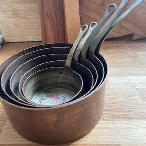 May include: A collection of five copper saucepans, each with a brass handle, nested together. The pans are of varying sizes, with the largest at the base. The copper has a warm, reddish-brown hue. The background features wooden chopping boards.