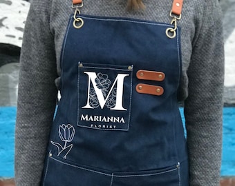 May include: A dark blue denim apron with white stitching, leather straps and a large pocket. The apron has the text "MARIANNA FLORIST" with a stylized "M" and a floral design. The apron is designed for use in a floral shop.