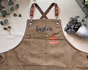 May include: Brown canvas apron with adjustable straps, a large pocket, and two smaller pockets. The apron has the name "Taylor" embroidered on the front and a simple line drawing of a tulip.