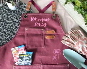 May include: A burgundy gardening apron with the text "Whoopsie Daisy" in pink script. The apron has two pockets, leather accents, and a floral design. Two seed packets and gardening gloves are also visible.