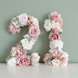 以下が含まれることがあります： 数字の21の形をしたフラワーアレンジメント。数字は、ピンク、白、ブラッシュカラーのバラやその他の花で構成され、緑の葉が付いています。アレンジメントは、くすんだ緑色の背景に配置されています。