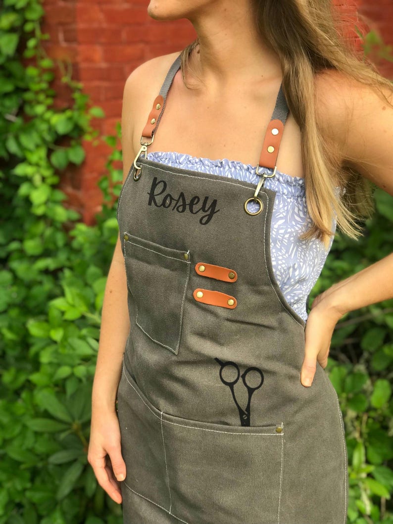 May include: A gray apron with the name "Rosey" printed on the front. The apron has pockets, leather accents, and a pair of scissors in one pocket. The straps are gray with brown leather accents.