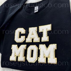Może przedstawiać: Czarna bluza z napisem "CAT MOM" w białych, złotych literach w stylu uniwersyteckim. Tekst jest wyśrodkowany na piersi. Nazwa marki "roseystudioco.com" jest wydrukowana na górze i na dole bluzy.
