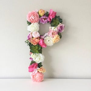 May include: A floral letter P made of pink, white and peach artificial flowers. The letter is surrounded by greenery.