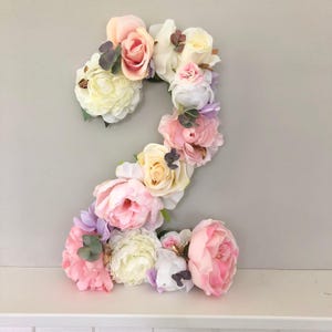May include: A floral arrangement in the shape of the number two. The number is composed of various pastel-colored flowers, including pink, white, and yellow roses and peonies. The flowers are arranged on a white surface against a light gray background.