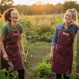 May include: Two women in a field of sunflowers, wearing burgundy aprons with leather straps. The aprons are personalised with the names "Grace" and "Mary". The women are smiling and laughing in a sunny outdoor setting, with a garden trowel visible.